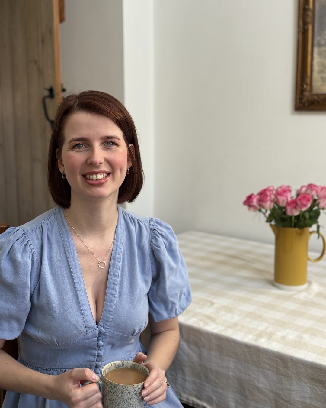 Emily smiling at her kitchen table with a mug of tea, a jug of pink roses beside her