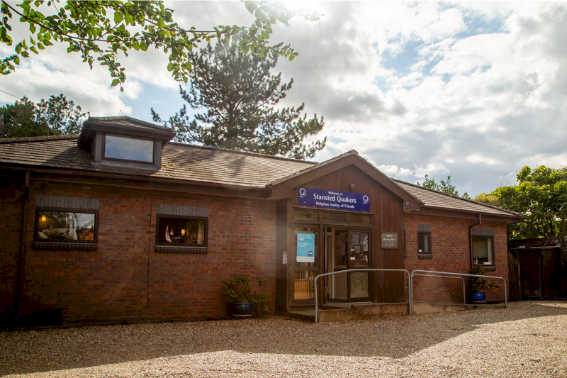 The Quaker Meeting House in Stansted on a sunny day: a single-storey red-brick building with a timber-clad porch, a Stansted Quakers sign, glass doors and a ramp with handrails.